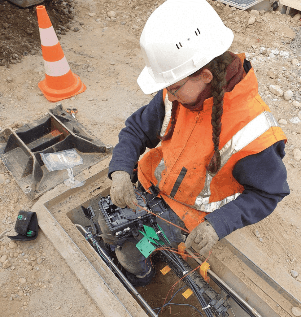Technicienne en veste orange et casque blanc raccordant des câbles fibre optique dans une chambre de tirage sur un chantier de déploiement télécom