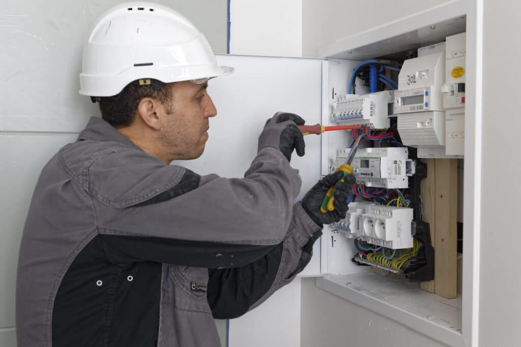 Technicien en combinaison grise et casque blanc câblant un tableau électrique résidentiel avec compteur communicant